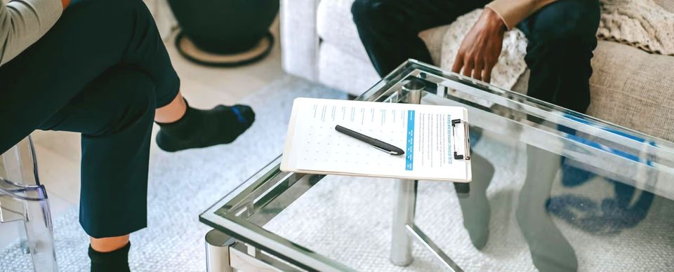 Two people sit on a sofa with a clipboard and pen on a glass table between them.